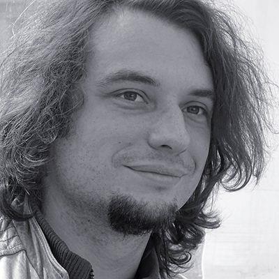 Porträt von Martin Fiedler mit schulterlangem Haar und Ziegenbart, leicht lächelnd und seitlich an der Kamera vorbeiblickend vor hellem, unscharfem Hintergrund